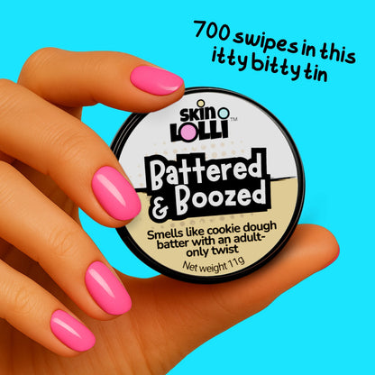 Hand holding tin of 'Battered & Boozed' cookie dough scented solid perfume by Skin Lolli on aqua blue background